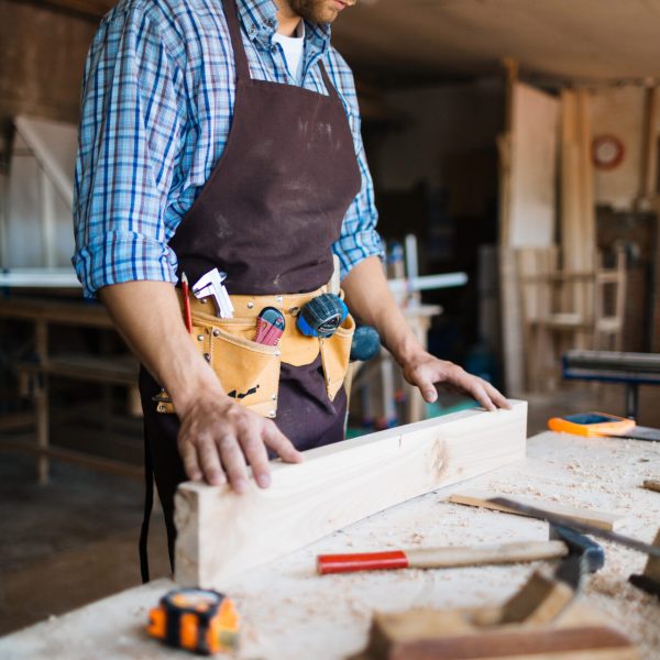 Bearded carpenter with pencil behind his ear checking surface of wooden board, table with shavings and hand tools on foreground